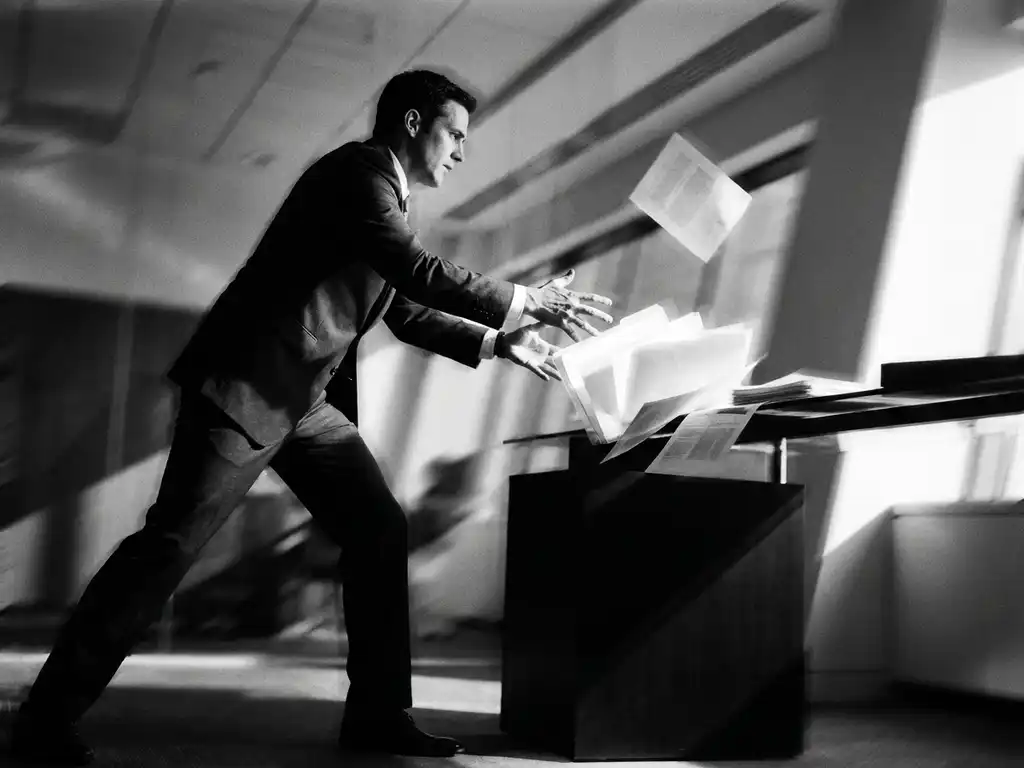 Zakenman in maatpak grijpt gloeiende documenten die van een modern bureau vallen, dramatische zijbelichting, zwart-wit filmstijl.